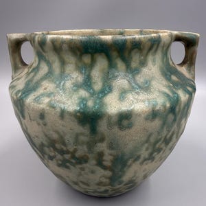 May include: A ceramic vessel with two side handles, featuring a textured, off-white base glaze overlaid with mottled, dripping patterns of teal and muted brown. The pot has a wide, rounded body and a thick, slightly flared rim opening.