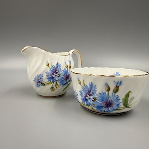 May include: A white porcelain creamer and sugar bowl set. Both pieces feature a blue floral design with gold trim. The creamer has a handle and spout, while the sugar bowl is open. The set is ideal for serving tea or coffee.