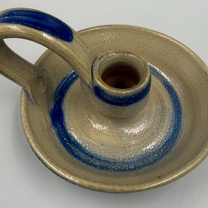 May include: A beige ceramic candle holder with a circular base and a single arched handle. The piece features a textured, speckled finish and is decorated with a vibrant blue painted stripe that circles the central candle cup and handle.