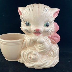 May include: A pink ceramic planter shaped like a cat with a pink bow. The cat has green eyes and a white face. The planter has a separate pot for planting.