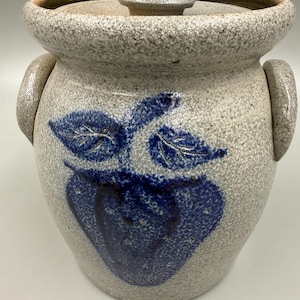 May include: A stoneware crock with a lid and two handles. The gray speckled pot features a blue apple design with leaves. The lid and handles are also gray. This vintage kitchenware is ideal for storage.