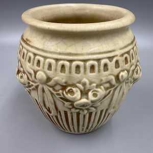 May include: Cream-colored ceramic pot with a wide rim and decorative relief patterns. The pot features a band of arched designs and floral motifs around the middle. Vertical lines and floral details adorn the lower portion of the pot.