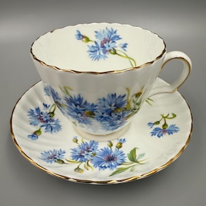 May include: White teacup and saucer set with a blue floral design. The teacup and saucer have a scalloped edge and gold trim. The blue flowers are cornflowers. The teacup has a handle.