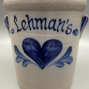 May include: A stoneware crock with a light beige speckled finish. The crock features a blue heart design with leaf accents and the word "Lehman's" in blue script. It has two small handles.
