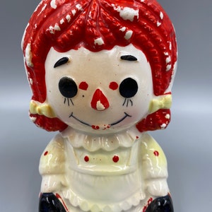 May include: A vintage ceramic Raggedy Ann piggy bank with red hair, a white dress with red polka dots, and black shoes.