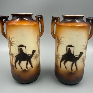 May include: A pair of ceramic vases featuring a gradient brown and cream finish. Each vase has two side handles and a central black silhouette graphic depicting a person riding a camel amidst palm trees, with dark brown rims and bases.