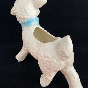May include: White ceramic lamb planter with a blue ribbon around its neck. The lamb is standing on all four legs and has a textured surface.