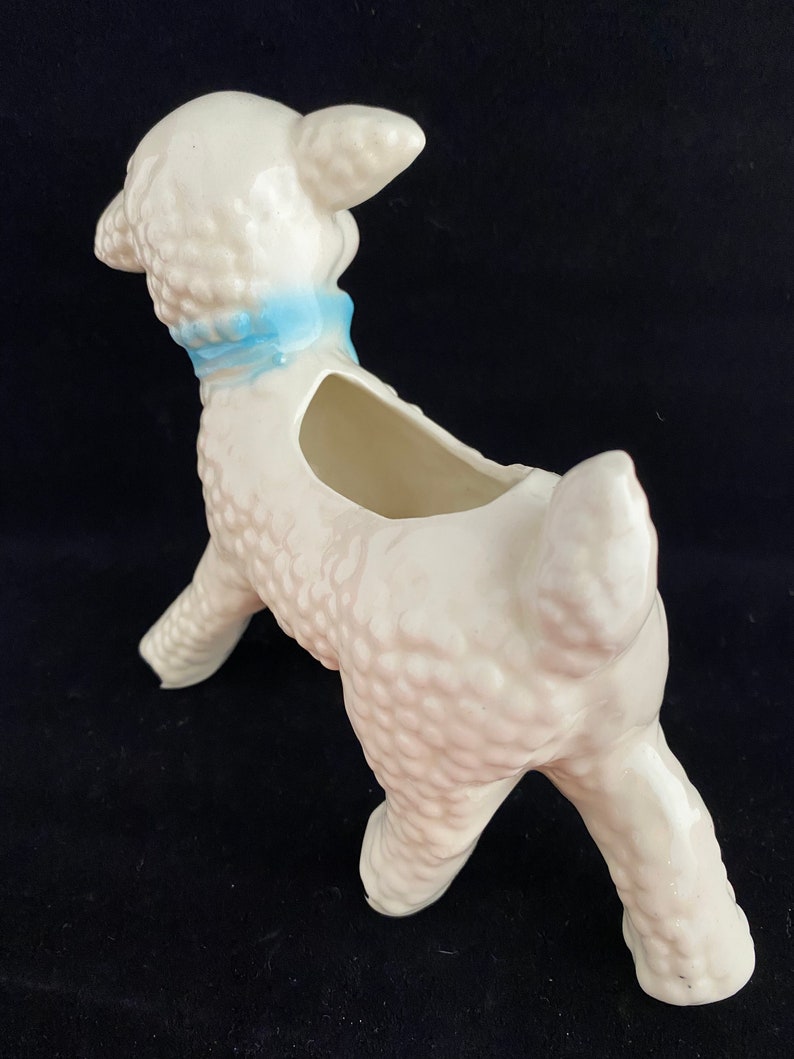 May include: White ceramic lamb planter with a blue ribbon around its neck. The lamb is standing on all four legs and has a textured surface.