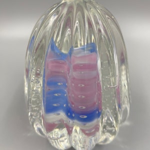 May include: A clear glass paperweight with a textured, ribbed surface. The paperweight contains a swirl of pink and blue glass with air bubbles.