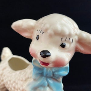 May include: A white ceramic lamb planter with a blue bow. The lamb has a pink nose and black eyes. The planter has a hole in the top for planting.