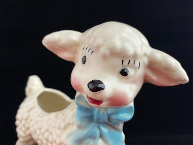 May include: A white ceramic lamb planter with a blue bow. The lamb has a pink nose and black eyes. The planter has a hole in the top for planting.