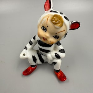May include: A vintage ceramic figurine of a seated figure in a black and white spotted outfit with red accents. The figurine has a smiling face, red shoes, and red-tipped ears. The figure has a yellow patch on its head.