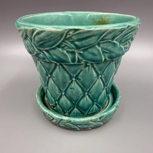 May include: A teal ceramic planter with a matching saucer. The planter features a quilted diamond pattern on the body and a leaf design around the rim. The saucer has a matching leaf design. The planter is approximately 7 inches tall.