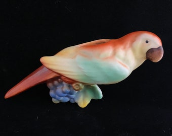 May include: A ceramic parrot figurine in shades of orange, yellow, and green, perched on a branch with blue grapes. The bird has a brown beak and a small black eye. The figurine is likely a decorative item.