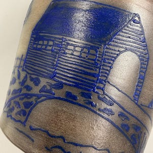 May include: A cylindrical ceramic vessel with a smooth, matte, light brown finish. The surface features a raised, vibrant blue relief design depicting a small house with a gabled roof, a stone bridge, and a fence, all rendered in bold lines.