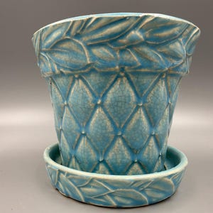 May include: A turquoise ceramic flower pot with a matching saucer. The pot features a diamond pattern and leaf-like detailing around the rim and on the saucer. The pot has a crackle glaze finish.