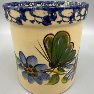 May include: A cylindrical ceramic planter with a cream-colored base, featuring a hand-painted green butterfly and blue flowers. The rim is decorated with a thick, mottled blue sponge-painted pattern. The vessel has a smooth, glossy finish.