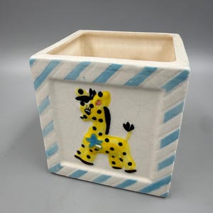 May include: A square ceramic planter featuring a white base with light blue diagonal stripes along the edges. The front panel displays a raised, hand-painted yellow giraffe with black spots, a black mane, and a small blue flower on its side.