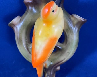 May include: A ceramic wall planter shaped like a bird perched on a branch. The bird is yellow and orange with a black beak. The branch is brown and green.