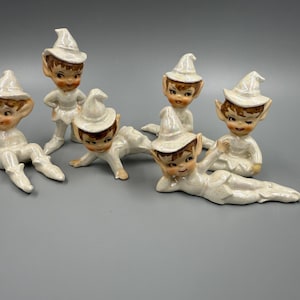 May include: A collection of six ceramic elf figurines. Each elf has a white hat and outfit, with brown hair and rosy cheeks. The elves are in various poses, some standing, crawling, or reclining.