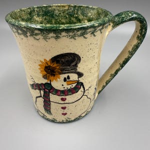 May include: Cream-colored ceramic mug with a hand-painted snowman design. The snowman wears a black hat, a sunflower, and a red and green scarf. The rim and handle are green.