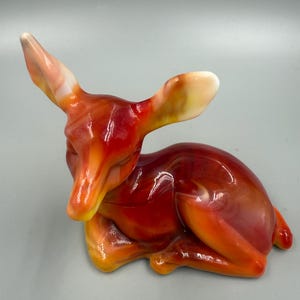May include: A decorative figurine of a deer in a resting pose. The deer is primarily red and orange with white accents on the ears. The figurine has a glossy finish and is likely made of glass or ceramic.