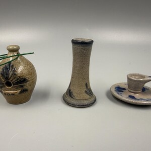 May include: Three ceramic pieces: a small, round birdhouse with a green wire handle, a slender vase, and a candle holder. All pieces are tan with blue floral designs. The candle holder has a small handle.