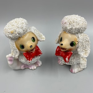 May include: Two white porcelain poodle figurines with red bows. The poodles have a fluffy, textured surface and green eyes.