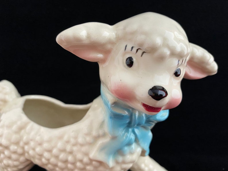 May include: A white ceramic planter shaped like a lamb with a blue bow. The lamb has pink cheeks and black eyes. The planter has a hole in the top for planting.