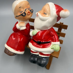 May include: Vintage ceramic Santa Claus and Mrs. Claus salt and pepper shakers. Santa wears a red suit with a black belt and boots, and Mrs. Claus wears a red dress. They are seated on a wooden bench. A charming Christmas decoration.