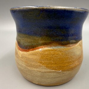 May include: A handmade ceramic mug with a unique, layered design. The top is a deep blue, transitioning to earthy tones of brown, orange, and yellow. The bottom is a light tan. The mug has a slightly flared rim and a rounded body.
