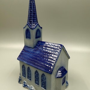 May include: A ceramic church figurine with a tall, pointed steeple and a blue roof. The building is primarily gray with blue accents, including the roof and the arched windows. The steeple has a textured, ribbed design.