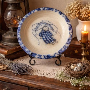 May include: A ceramic plate with a blue and white angel design. The plate has a blue rim and is displayed on a black metal stand. The plate is on a wooden table with a lit candle and other decorative items.