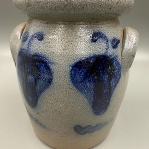 May include: A stoneware crock with a light gray speckled finish and dark blue leaf-like designs. The antique jar has two side handles and a lid. The ceramic container is a vintage kitchenware item.