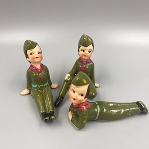 May include: Three ceramic figurines of boy scouts in green uniforms with red and blue accents. The figurines are sitting and lying down.