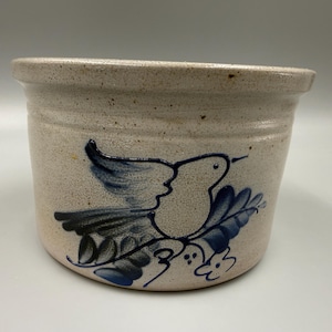 May include: A beige ceramic pot with a blue bird and leaf design. The pot has a rounded shape with a slightly flared rim. The bird is depicted in flight, with detailed wing and tail feathers.
