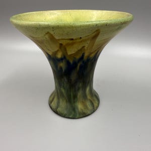 May include: A ceramic vase with a flared top and a rounded base. The vase features a color gradient, transitioning from a light green rim to a dark green base, with brown and blue accents. The vase has a unique, organic shape.