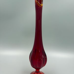 May include: A tall, slender glass vase featuring a deep red body with a bulbous, ribbed base and a flared, yellow-tinted rim. The vase stands on a circular pedestal foot and displays a clear, elongated neck that tapers toward the top opening.