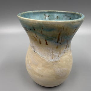 May include: A handmade ceramic vase with a wide, flared top and a rounded base. The vase features a gradient of colors, transitioning from a light blue rim to a tan and white body. The interior is a light blue color.