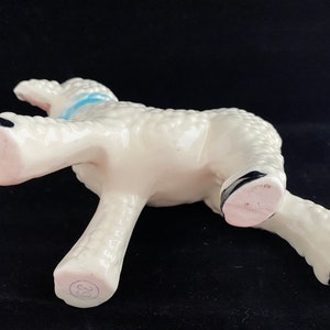 May include: White ceramic lamb figurine with a blue ribbon around its neck. The lamb is standing on all four legs and has a textured surface. The figurine is marked with the number "38" on the bottom.