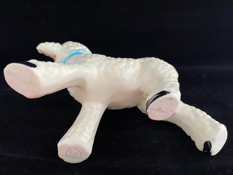 May include: White ceramic lamb figurine with a blue ribbon around its neck. The lamb is standing on all four legs and has a textured surface. The figurine is marked with the number "38" on the bottom.