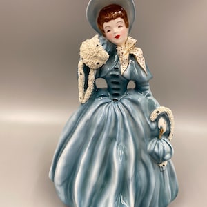 May include: A vintage ceramic figurine of a woman in a blue dress and white hat. She is holding a white beaded bag and has a white bow around her neck.