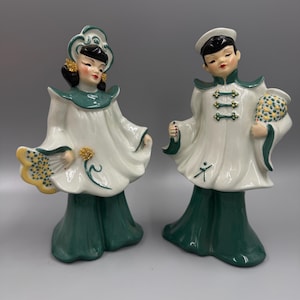 May include: A pair of vintage ceramic figurines depicting figures in traditional Asian-inspired attire. The figurines feature white and green robes, with detailed facial features and accessories. One holds a fan, the other a pouch.
