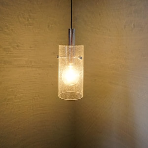 May include: A modern pendant light with a cylindrical glass shade. The light fixture has a chrome finish and a single bulb, creating a warm glow. The design is minimalist and suitable for various interior styles.