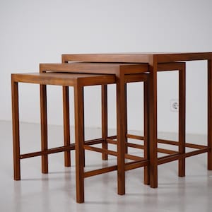 Könnte beinhalten: Ein Set aus drei Satztischen aus mittelbraunem Holz. Jeder Tisch verfügt über eine rechteckige Platte und vier gerade Beine, die durch horizontale Stützstreben nahe der Basis verbunden sind. Die Tische sind treppenförmig angeordnet.