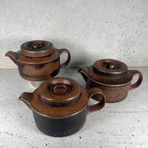 Op de afbeelding: Drie keramische theepotten met deksels. De theepotten zijn bruin en zwart met een textuurafwerking. Elke theepot heeft een handvat en een tuit. De theepotten zijn geplaatst op een grijze achtergrond.