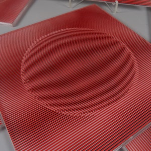 Könnte beinhalten: Rote quadratische Fliese mit einer kreisförmigen Vertiefung in der Mitte. Die Fliese hat eine strukturierte Oberfläche mit dünnen, parallelen Linien.