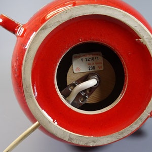 May include: A red ceramic lamp base with a glossy finish. The lamp has a round shape with two small handles and a white rim. The bottom reveals the electrical components and a label with text. The lamp is designed to hold a light bulb.