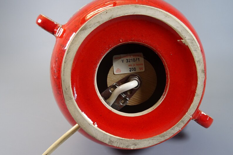 May include: A red ceramic lamp base with a glossy finish. The lamp has a round shape with two small handles and a white rim. The bottom reveals the electrical components and a label with text. The lamp is designed to hold a light bulb.