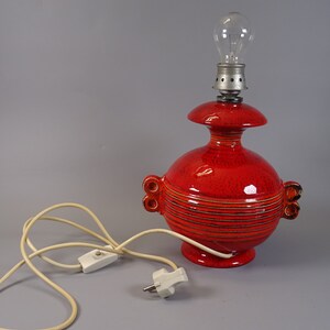 May include: A red ceramic table lamp with a bulb. The lamp has a round base and body with horizontal lines and two small handles. The lamp is connected to a white cord with a switch and plug.